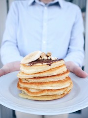 Man is holding plate with freshly made homemade pancakes with chocolate nougat, grounded peanuts and walnut, sliced banana. Sweet breakfast, food for freakfast. Gluten free pancakes. Closeup