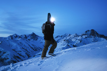 Brave explorer with headlamp and backpack and a snowboard behind his back climb on great snowy mountain at night. Traveler wearing ski wear. Amazing view on winter ridge © leonidkos
