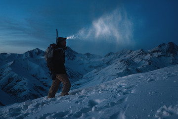 Brave man with headlamp, backpack and a snowboard behind his back climb night on snowy mountain. Man commit ski tour in high ridge. Backcountry. Great view on winter mountains