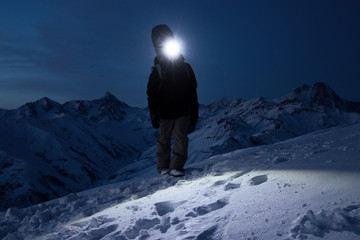 Professional tourist climb on snowy mountain at night and lights the way with a headlamp. Snowboarder walking in front of amazing winter mountains view with backpack and a snowboard behind his back © leonidkos