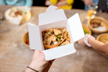 Holding a box with asian food indoors with table full of food on the background