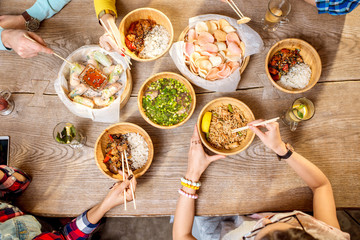 Top view on the table full of different asian meals served in the wooden plates and young people eating with sticks