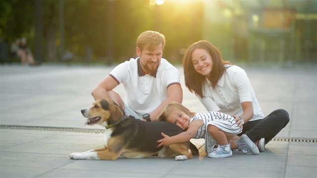 Lovely Family Hugging Each Other Sitting on the Ground During Summer Sunset in the City. Cute Daughter Hugs Beagle Dog, Young Husband Kisses His Wife.