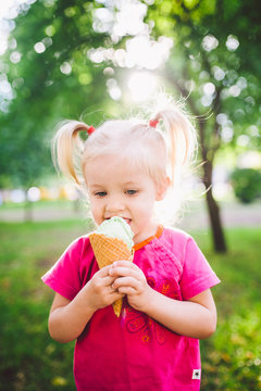 little funny girl blonde eating sweet blue ice cream in a waffle cup on a green summer background in the park. smeared her face and cheeks and laughs. Dressed in bright stylish clothes
