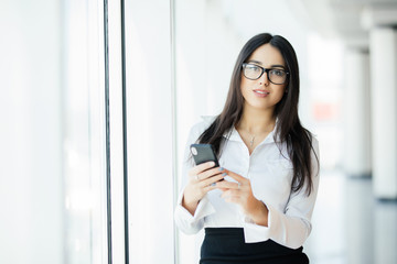 Obraz premium Portrait of a young business woman in glasses typing text phone against panoramic windows. Business concept