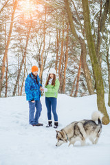 Fototapeta na wymiar Sporty cute hipster couple in bright winter sportswear having fun in winter park with their friend husky alaskan malamute dog on a day talking with each other and smiling.