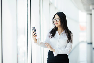 Portrait of a young business woman in glasses typing text phone against panoramic windows. Business concept