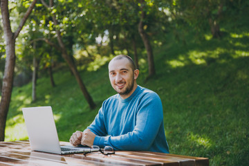 Young successful smart man businessman or student in casual blue shirt glasses sitting at table with mobile phone in city park using laptop working outdoors on green nature. Mobile Office concept.