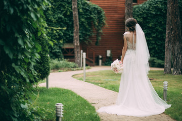 Fototapeta premium The bride stands with her back in the wedding dress with a bouquet in her hand on the alley in the nature.