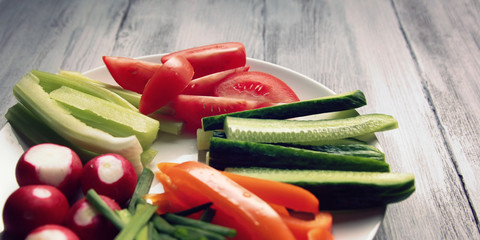 White plate with vegetables for a vegetarian salad. White wooden kitchen table. Radishes, tomatoes, celery, bell pepper, onion and cucumber. Close up. Wide photo.