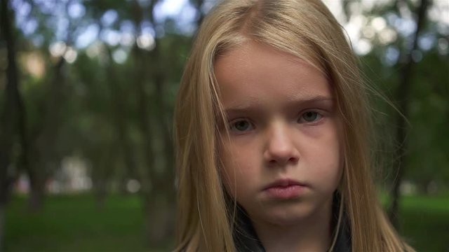 Serious little girl with light hair looking at camera while standing in a park on a cloudy summer day. Handheld slow motion close up shot