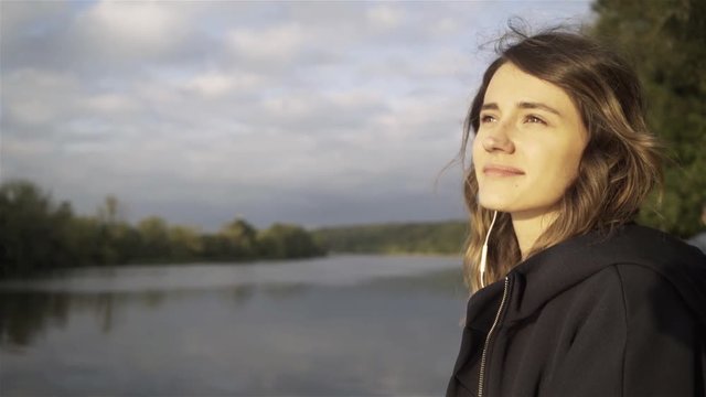 Cheerful young woman wearing a black hoodie listening to the music and standing on a river bank. Tracking real time establishing shot