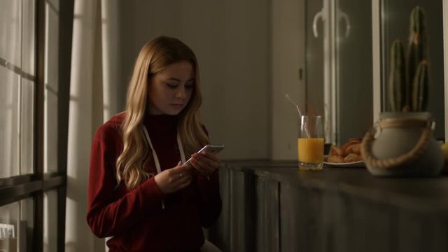 Stunning long blonde hair young woman connecting to social networks with smart phone and drinking juice while spending leisure in domestic interior at night. Girl reading social media threads on phone