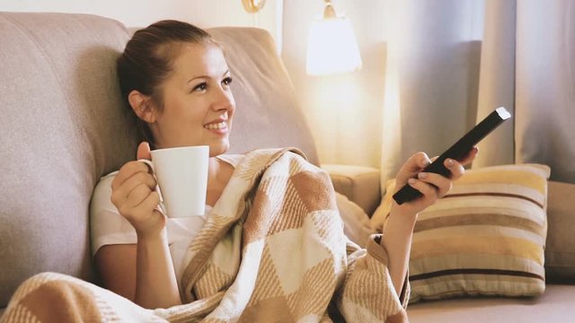 Young female is watching TV and sitting with tea at home.