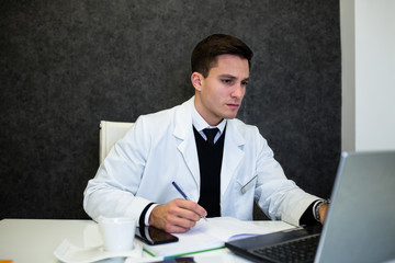 Young and handsome doctor sitting in his office and seriously looking at laptop.