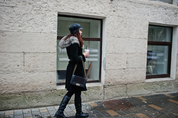 Curly mexican girl in leather cap and plastic cup of coffee at hand walking at streets of city.