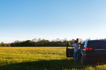 The young couple stands next to the SUV at the door and points to the distance of the hand on the summer field with the door open. A man and a girl near the car enjoy the landscape. Place for text.