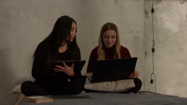 Smart diverse girls studying together using laptop pc and digital tablet while sitting cross-legged on the bed at home. Positive female students working with modern technology devices and chatting.