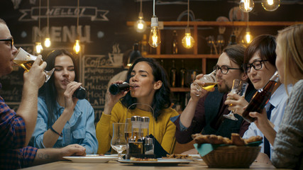 Diverse Group of Friends Celebrate with a Toast and Clink Raised Glasses with Various Drinks in Celebration. Beautiful Young People Have Fun in the Stylish Bar/ Restaurant.