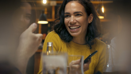 Beautiful Hispanic Woman Uses Smartphone While Talking and Having Fun with Her Friends in the Bar. They Laugh, Joke, Drink in Stylish Hipster Bar Establishment.