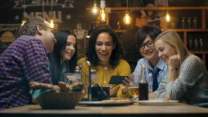 Beautiful Hispanic Woman Shows Interesting Stuff on Her Smartphone to Her Friends while They Have Good Time in Bar. They Laugh, Joke, Drink in Stylish Hipster Bar Establishment.