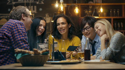 Beautiful Hispanic Woman Shows Interesting Stuff on Her Smartphone to Her Friends while They Have Good Time in Bar. They Laugh, Joke, Drink in Stylish Hipster Bar Establishment.