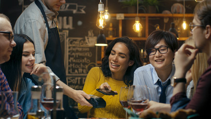 In the Bar Waiter Holds Credit Card Payment Machine and Beautiful Woman Pays for Her Order with Contactless Mobile Phone Payments System. She Has Good Time with Her Friends.