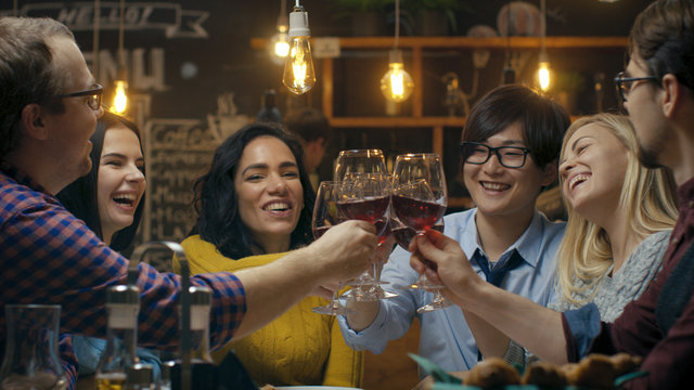 Diverse Group of Friends Celebrate with a Toast and Clink Raised Wine Glasses in Celebration. Beautiful Young People Have Fun in the Stylish Bar/ Restaurant.