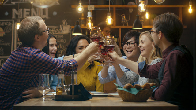 Diverse Group of Friends Celebrate with a Toast and Clink Raised Wine Glasses in Celebration. Beautiful Young People Have Fun in the Stylish Bar/ Restaurant.