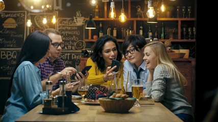 In the Bar/ Restaurant Birthday Party Celebration in Progress Beautiful Young People Show Their Mobile Phone Screens and Have Great Fun.