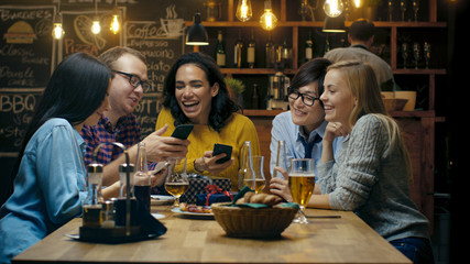 In the Bar/ Restaurant Birthday Party Celebration in Progress Beautiful Young People Show Their Mobile Phone Screens and Have Great Fun.