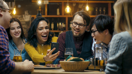 Beautiful Hispanic Woman Shows Interesting Stuff on Her Smartphone to Her Friends while They Have Good Time in Bar. They Laugh, Joke, Drink in Stylish Hipster Bar Establishment.