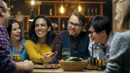 Beautiful Hispanic Woman Shows Interesting Stuff on Her Smartphone to Her Friends while They Have Good Time in Bar. They Laugh, Joke, Drink in Stylish Hipster Bar Establishment.