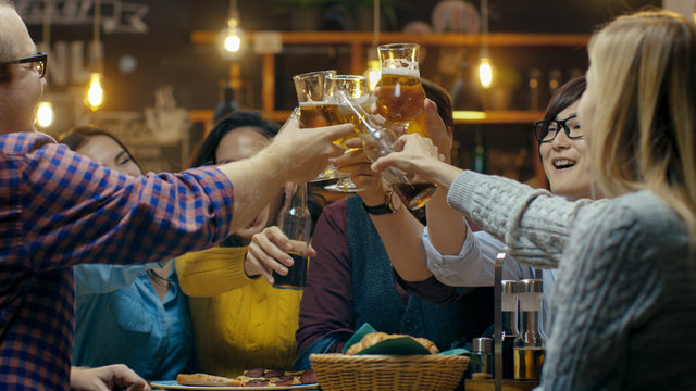 Diverse Group of Friends Make a Toast and Clink Raised Glasses with Various Drinks in Celebration. Beautiful Young People Have Fun in the Stylish Bar/ Restaurant.