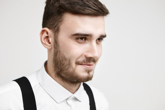 Half profile picture of friendly looking fashionable young male with mustache and beard smiling thoughtfully as he reminds some funny story or joke, posing in studio wearing white formal shirt