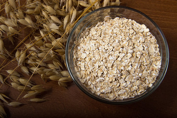 Oat ears stems and oat flakes in a bowl on a dark brown wood background. Top view. oat flakes small size grind. Close up. Useful fiber-rich product. Dietary breakfast from healthy foods
