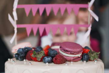 On the children's birthday cake there are raspberry berries, blueberries and strawberries, purple and pink macaroons, decorative pink flags and ribbons. Close up.