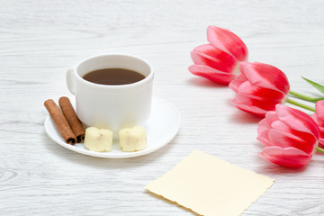 Three pink tulips, mug of coffee and cinamon, light wooden background.