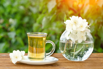Transparent mug of tea and a vase with jasmine. Greens on the background, sunlight