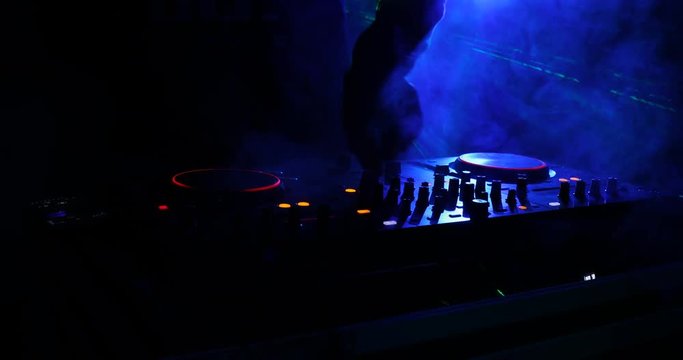 DJ Spinning, Mixing, and Scratching in a Night Club, Hands of dj tweak various track controls on dj's deck, strobe lights and fog, selective focus, close up. Slider Shoot