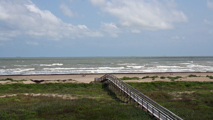 Boardwalk through green marsh grass leading to the sand and surf of Galveston Beach, Texas