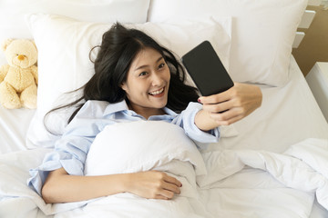 Top view happy smiling girl using smart phone for selfie on the bed. Beautiful young woman self-portrait with mobile camera lying on white bed. Concept lifestyle technology