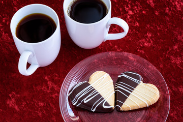Two heart shaped shortbread cookies, dipped in chocolate, on a clear glass plate on a luxurious red background, with two white cups of tea/coffee
