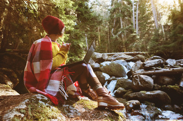 Naklejka na ściany i meble A toned portrait of a freelancer hipster girl with glasses and a stylish cap dressed in a blanket with a laptop kneeling sitting on a rock in a coniferous forest next to a stormy mountain river in