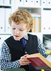 Young curly Caucasian boy reading book in library.