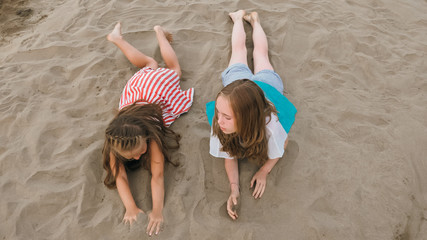 Two teenage are lie on a sandy beach near the sea. Girl playing, talk to each other. Sisters are dressed in dresses and sunglasses. Children have real emotions of happiness.