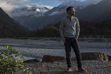 Retrato de hombre joven frente a paisaje de picos de montañas nevados y verdes con cielo nublado en Nueva Zelanda