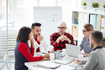 Group of three men and two women in smart casual gathered by table in office for meeting