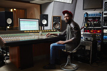 Young African-american rapper in baseball cap, jeans and leather jacket working by switchboard in studio - obrazy, fototapety, plakaty