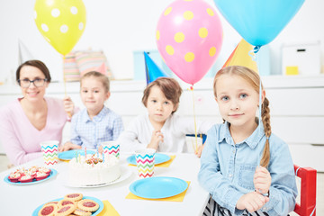 Cute child with pink balloon, her friends and teacher gathered by festive table at birthday party